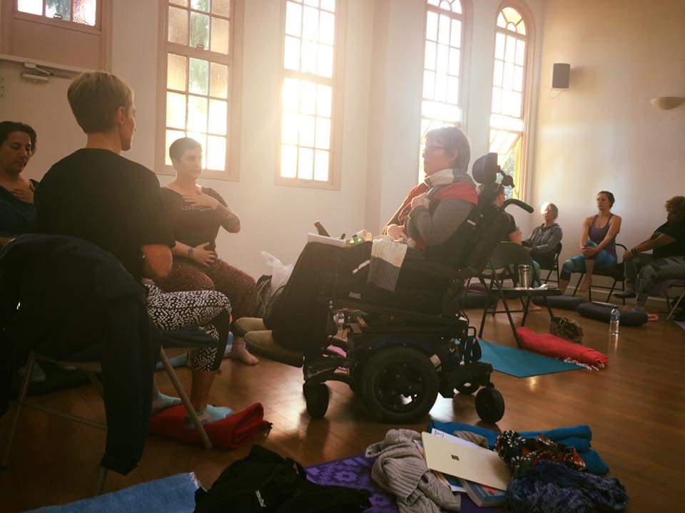 A yoga class with a group of four people. One woman  is in a power wheelchair, tilted back slightly