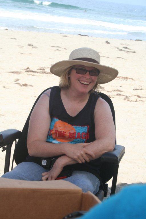 A white woman sitting in a wheelchair on the beach wearing glasses and a straw hat