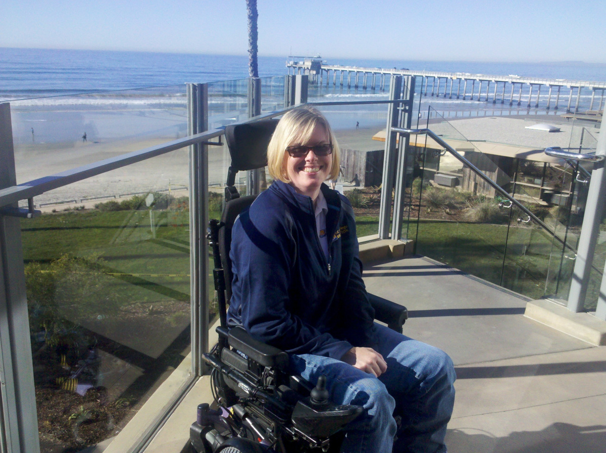 A white woman sitting in a black power wheelchair sitting on a deck with clear walls and  the ocean behind her