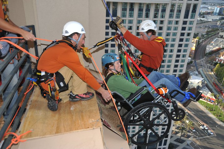 Two men helping a woman in a wheelchair rappel down a building