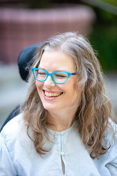 A white woman smiling, head turned to her right with dark blonde curly hair below her shoulders, wearing teal glasses and light blue shirt