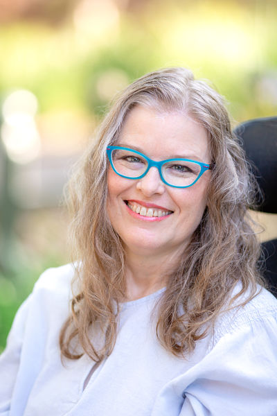 A white woman with dark blonde curly hair below her shoulders, wearing teal glasses and light blue shirt