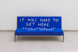 A blue bench with white text reads: IT WAS HARD TO GET HERE. REST HERE IF YOU AGREE.—inviting reflection on criptime—against a plain white wall on a tiled floor.
